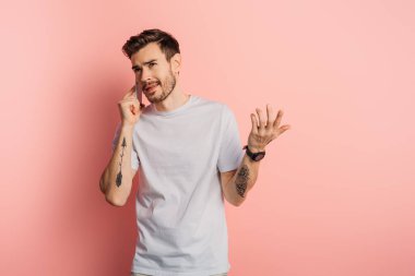Pembe arka planda akıllı telefonla konuşurken omuz silkme hareketi yapan kafası karışmış genç bir adam.