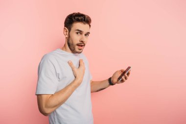 Şok olmuş genç adam pembe arka planda akıllı telefonu tutarken göğsüne dokunuyor.