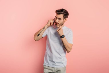 thoughtful young man touching chin while talking on smartphone on pink background clipart