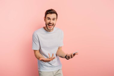 Pembe arka planda akıllı telefonu tutarken etkileyici bir jest yapan genç adam.