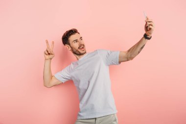 Heyecanlı genç adam pembe arka planda akıllı telefondan selfie çekerken zafer jesti yapıyor.