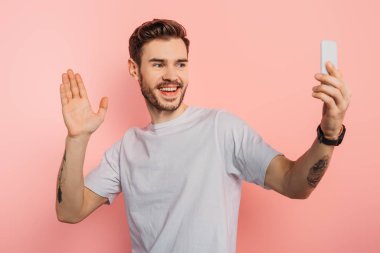 heyecanlı genç adam pembe arka planda akıllı telefondan görüntülü konuşma yaparken el sallıyor.