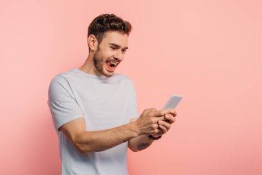 Heyecanlı genç adam pembe arka planda akıllı telefondan sohbet ediyor.