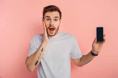 Pembe arka planda boş ekranlı akıllı telefon gösterilirken, şok olmuş bir adam yüzüne dokunuyor.