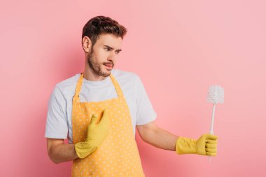 Önlüklü kafası karışmış genç bir adam ve pembe arka plandaki tuvalet fırçasına bakarken kendini gösteren lastik eldivenler.