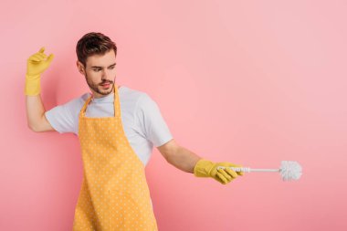 Önlüklü ve lastik eldivenli bir adam pembe arka planda tuvalet fırçasıyla eskrimi taklit ediyor.