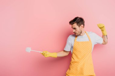 Önlüklü ve lastik eldivenli genç adam pembe arka planda tuvalet fırçasıyla eskrimi taklit ediyor.