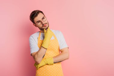 Önlüklü ve lastik eldivenli düşünceli genç adam çeneye dokunuyor ve pembe arka plana bakıyor.
