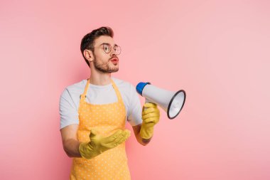 Pembe arka planda kolları açık dururken elinde megafon tutan önlüklü ve lastik eldivenli hoşnutsuz genç adam.