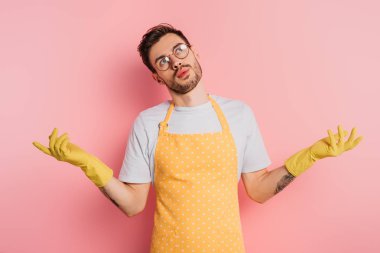 Pembe arka planda omuz silkme hareketi gösteren önlüklü ve lastik eldivenli kafası karışmış genç bir adam.