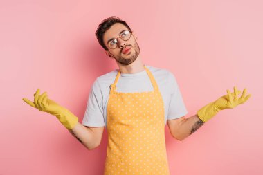 confused young man in apron and rubber gloves showing shrug gesture on pink background clipart