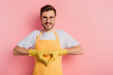 Pembe arka planda kalp hareketi gösteren önlüklü ve lastik eldivenli mutlu genç adam.