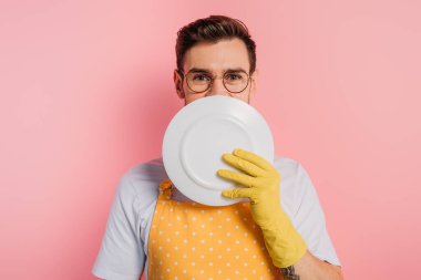 Önlüklü neşeli genç bir adam ve pembe arka planda temiz bir plaka ile yüzünü kaplayan lastik eldivenler
