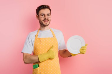 Önlüklü, lastik eldivenli, elinde temiz bir tabak tutan ve pembe arka planda baş parmağını gösteren mutlu bir genç.