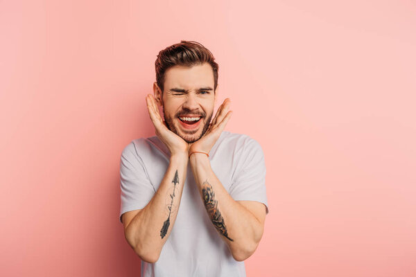 sly young man with hands near face smiling and winking on pink background