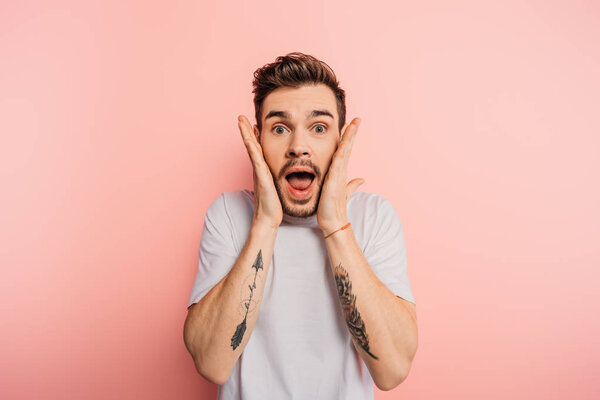 shocked young man with open mouth and hands near face looking at camera on pink background