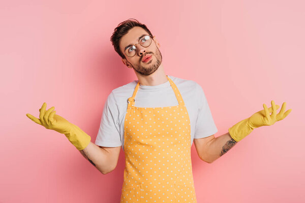 confused young man in apron and rubber gloves showing shrug gesture on pink background