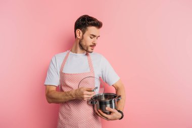 Önlüklü ciddi bir genç adam pembe arka planda tencereyi araştırıyor. 