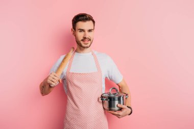 Önlüklü neşeli genç adam elinde tencere ve pembe arka planda merdaneyle 