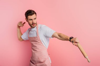 concentrated young man in apron imitating playing baseball with rolling pin on pink background clipart