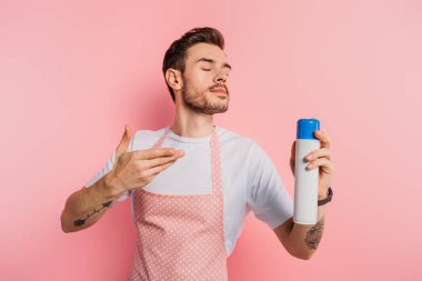 pleased young man in apron enjoying smell of air freshener on pink background clipart