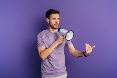 serious young man showing come here gesture while holding megaphone on purple background clipart