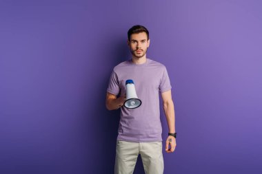 serious young man holding megaphone and looking at camera on purple background clipart