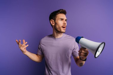 shocked young man holding megaphone and looking away on purple background clipart