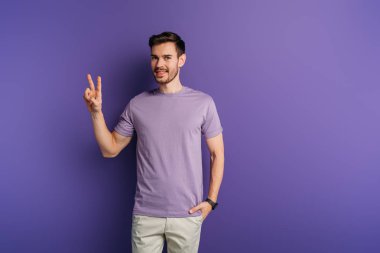 smiling young man showing victory gesture while holding hand in pocket on purple background clipart
