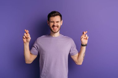 excited young man holding crossed fingers while standing with closed eyes on purple background clipart