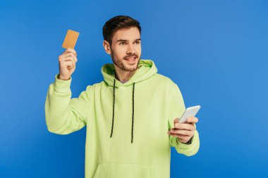 Akıllı telefon ve kredi kartı tutan neşeli genç adam mavi ışıkta gözlerden uzaklaşıyor.