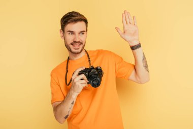 Sarı arka planda el sallayan dijital kameralı yakışıklı, neşeli fotoğrafçı.