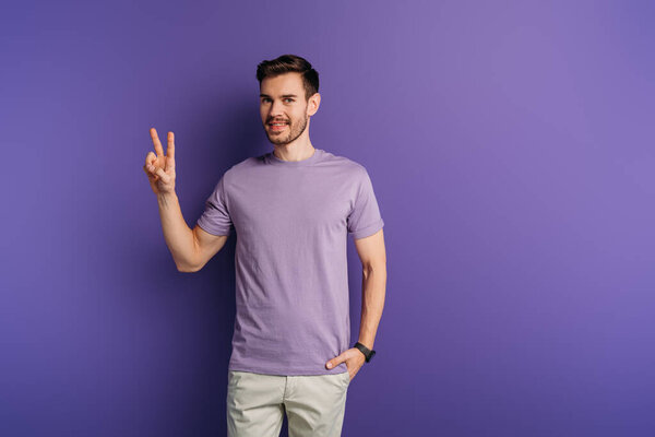 улыбающийся молодой человек показывает победный жест, держа руку в кармане на фиолетовом фоне
