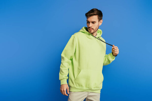 Красивый молодой человек трогает шнурки на толстовке, позируя изолированным на голубом
