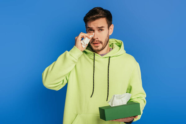 upset young man crying and wiping tears with paper napkin isolated on blue