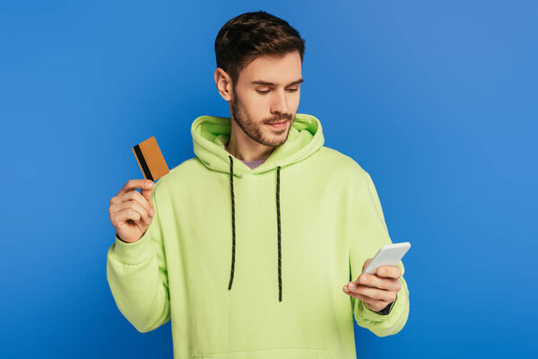attentive young man using smartphone while holding credit card isolated on blue