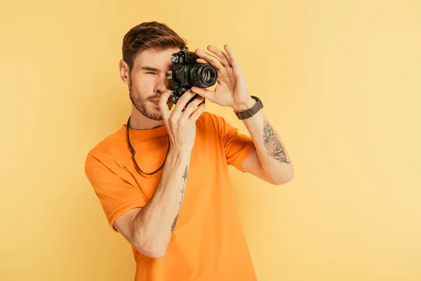 attentive handsome photographer taking picture with digital camera isolated on yellow
