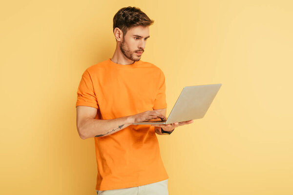 attentive young man in orange t-shirt using laptop on yellow background