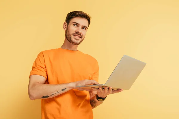 Smiling Dreamy Man Looking Away While Holding Laptop Yellow Background — Stock Photo, Image