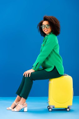 Gülümseyen Afrikalı Amerikalı kadın mavi arka planda seyahat çantasında oturuyor. 