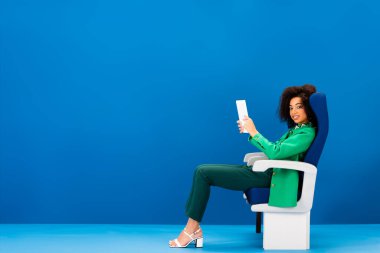 Gülümseyen Afrikalı Amerikalı kadın koltukta oturuyor ve elinde mavi arka planda dijital tablet var. 