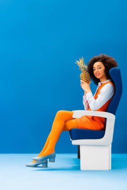 Gülümseyen Afrikalı Amerikalı retro elbiseli koltukta oturuyor ve mavi arka planda ananas tutuyor. 
