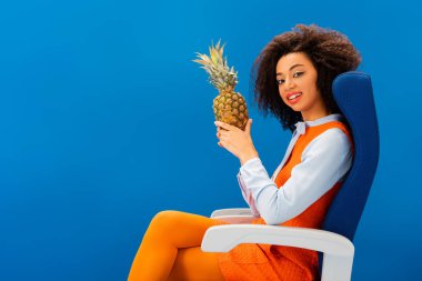 Gülümseyen Afrikalı Amerikalı retro elbiseli koltukta oturuyor ve mavi üzerinde ananas tutuyor. 