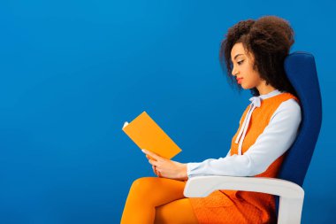 Eski moda elbiseli Afro-Amerikan 'ın yan görünüşü koltukta oturup mavi üzerine izole edilmiş kitap okuması. 