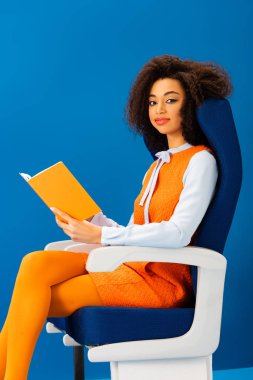 Gülümseyen Afrikalı Amerikalı retro elbiseler içinde kitap tutuyor ve mavi koltukta tek başına oturuyor. 