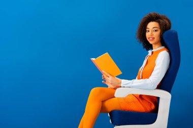 Gülümseyen Afrikalı Amerikalı retro elbiseler içinde kitap tutuyor ve mavi koltukta tek başına oturuyor. 