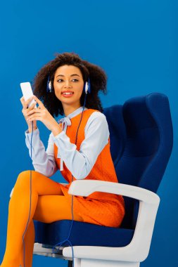 Gülümseyen Afrikalı Amerikalı retro elbiseli müzik dinliyor ve mavi üzerine izole edilmiş akıllı telefon kullanıyor. 