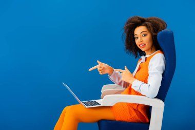 Gülümseyen Afro-Amerikan koltukta oturuyor ve mavi üzerine izole edilmiş dizüstü bilgisayarını işaret ediyor. 
