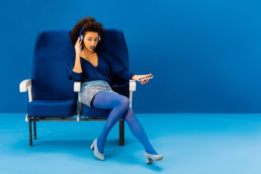 Afro-Amerikan koltukta oturmuş, müzik dinliyor ve mavi arka planda akıllı telefon tutuyor. 