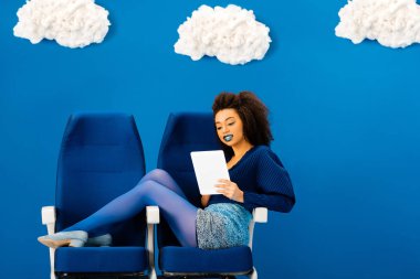 Gülümseyen Afro-Amerikan koltuklarda oturuyor ve bulutlu mavi arka planda dijital tablet kullanıyor. 
